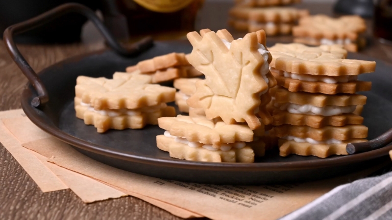 Homemade maple leaf cookies with creamy filling stacked neatly on a dark tray, ready to serve
