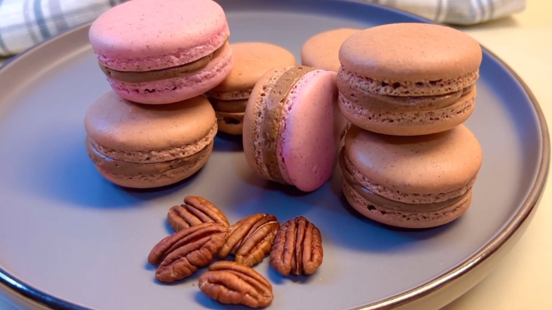 Pecan maple macarons with pink and brown shells stacked on a plate beside a few whole pecans